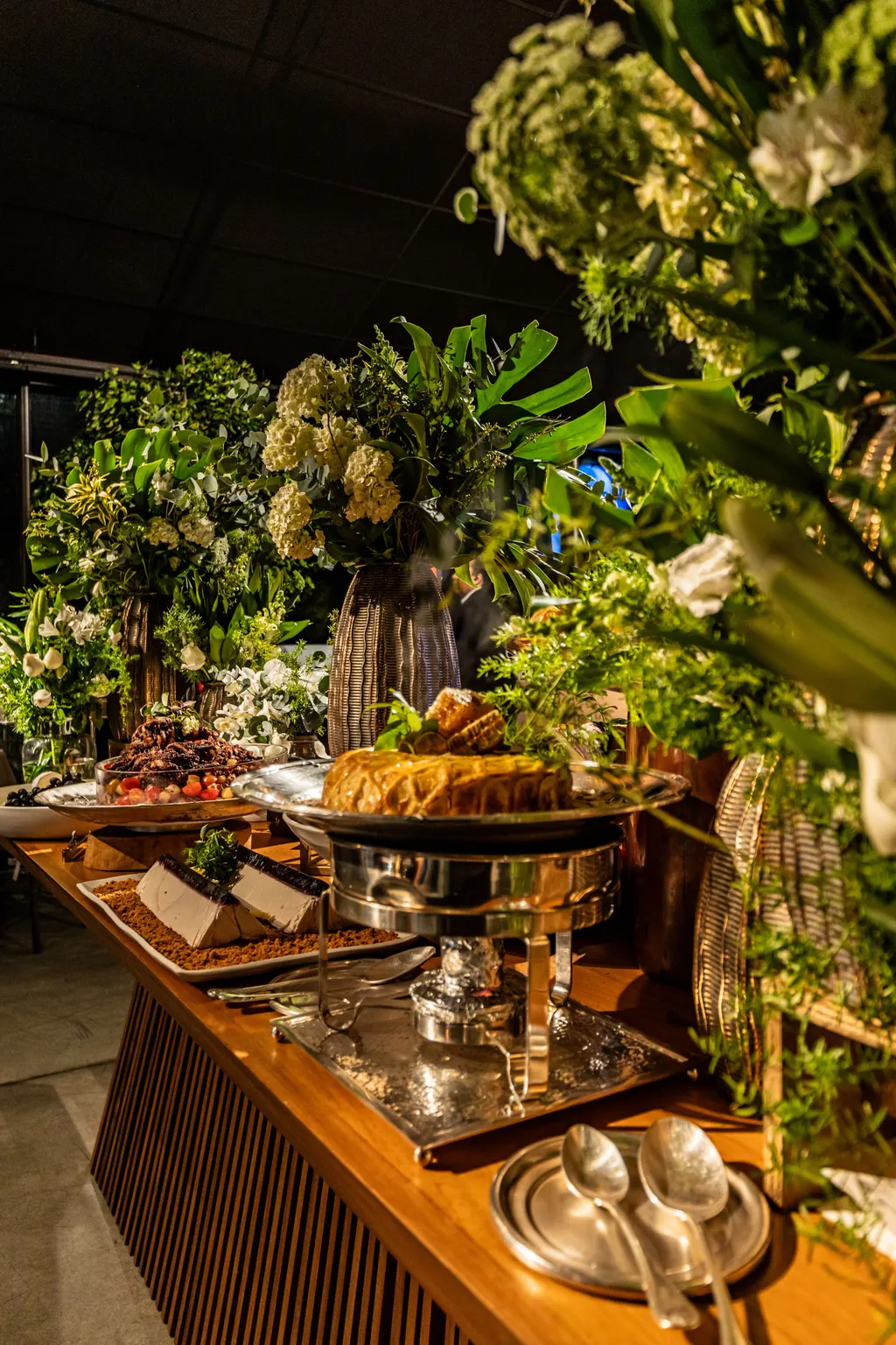 Mesa de buffet em madeira com réchaud de prata e arranjos de folhagens