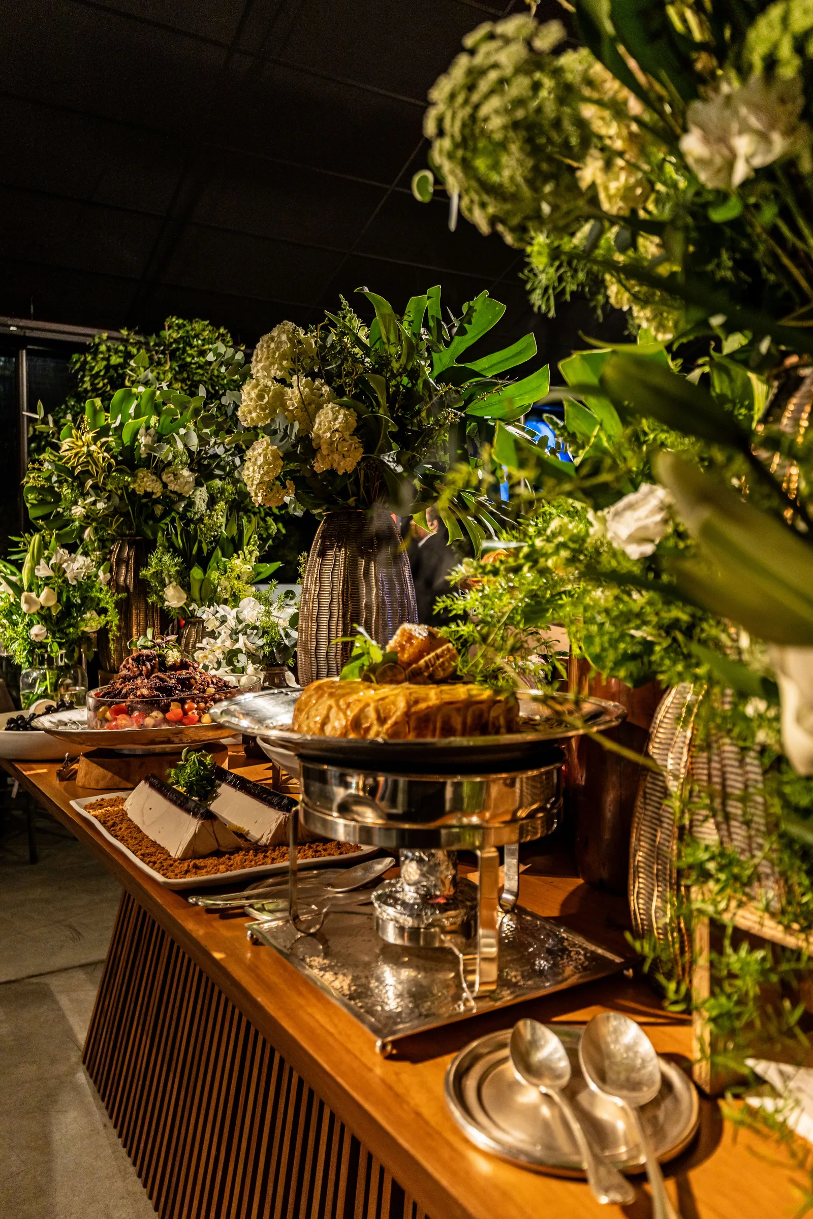Mesa de buffet em madeira com réchaud de prata e arranjos de folhagens em evento noturno