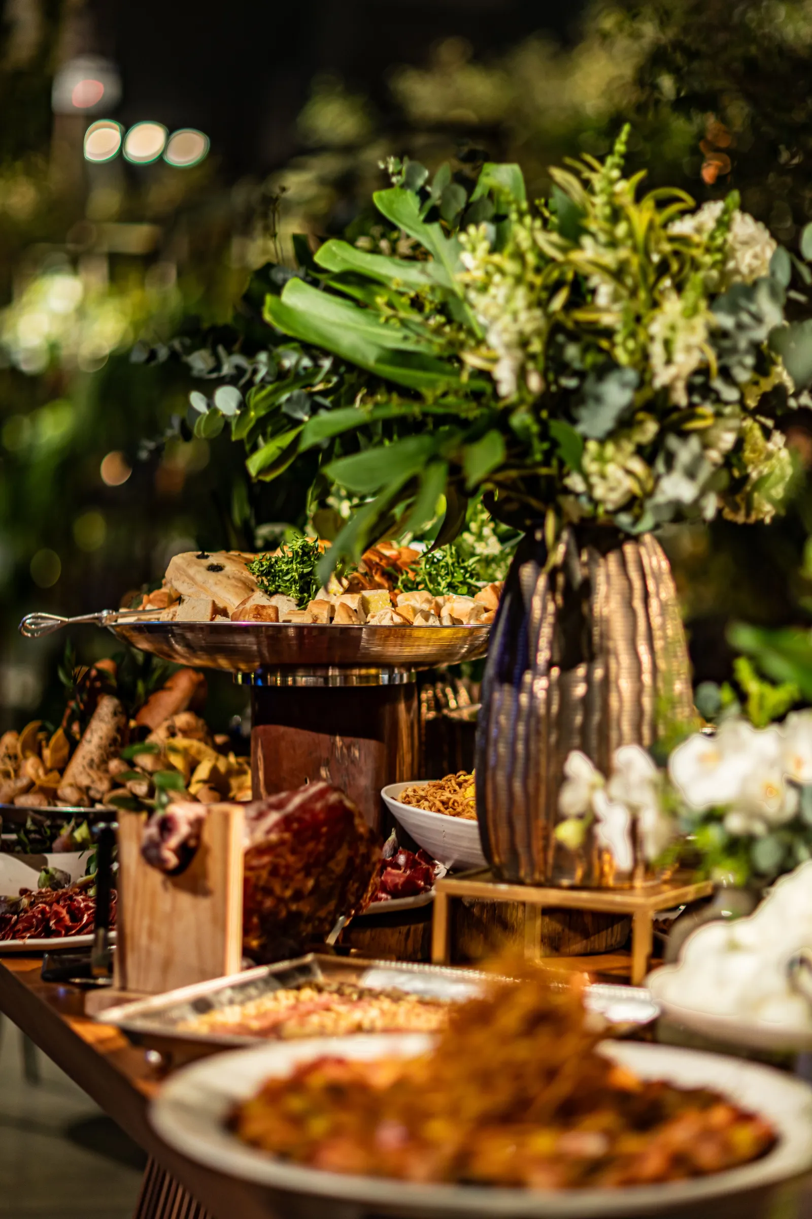 Travessa de prata com pães e antepastos diante de arranjo de folhagens e flores brancas