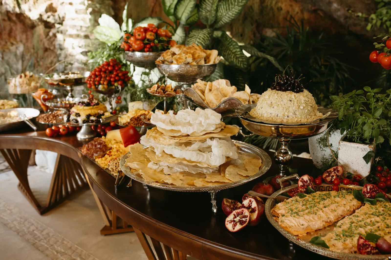 Mesa de buffet em madeira curva com travessas de prata, frutas e folhagens ao fundo